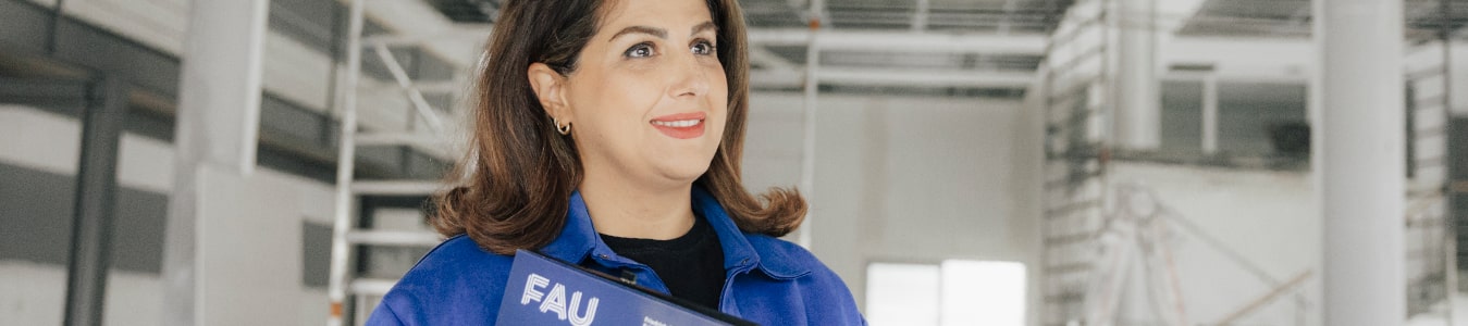 Weiblich gelesene Person steht auf einer Indoor-Baustelle. Sie trägt eine Mappe mit dem Logo der Friedrich-Alexander-Universität Erlangen-Nürnberg und lächelt.