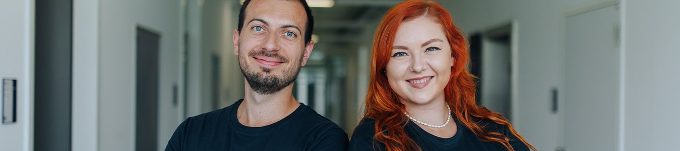 Eine männlich und eine weiblich gelesene Person lächeln in die Kamera. Beide tragen dunkelbaue T-Shirts. Die rechts stehende Person hat leuchtend rote Haare und trägt eine Perlenkette.