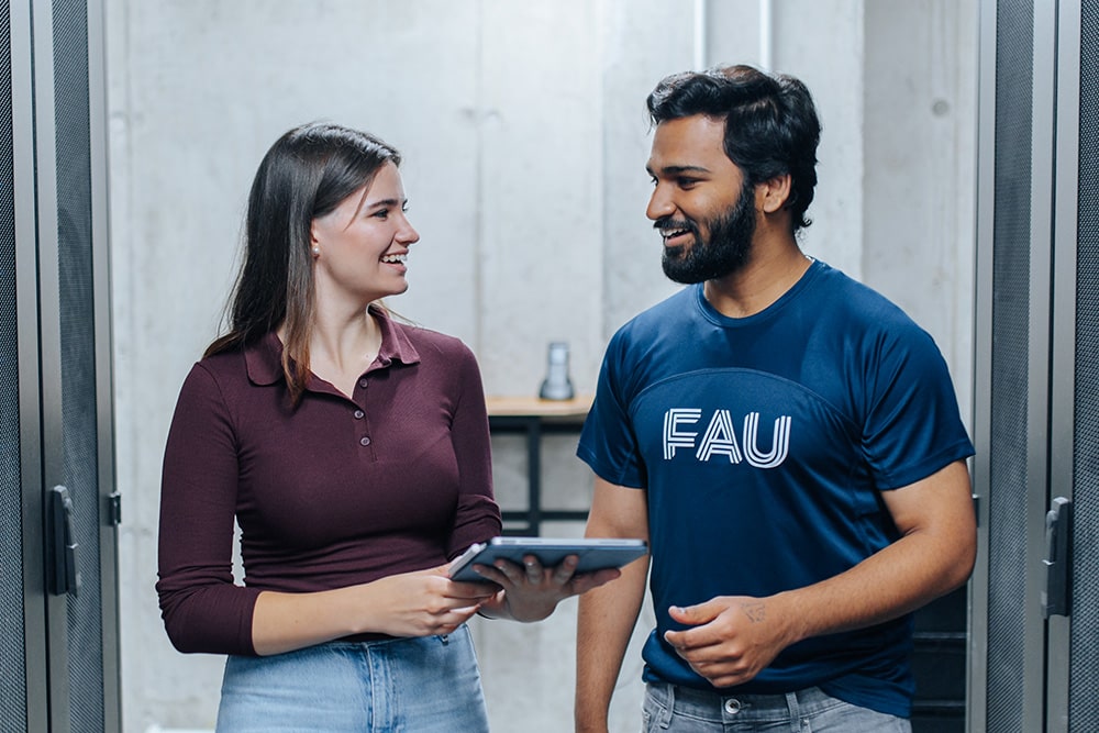 Zwei Personen stehen in einem Serverraum und sind in ein Gespräch vertieft. Die links stehende Person hält ein Tablet in der Hand, die rechts stehende Person trägt ein blaues T-Shirt mit dem Logo der Friedrich-Alexander-Universität Erlangen-Nürnberg.