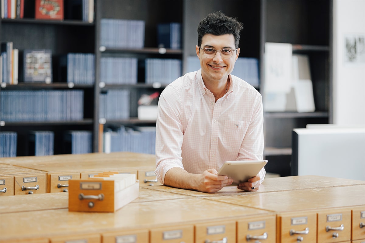 Männlich gelesene Person steht vor einem Regal mit blauen Buchbänden. Die Person steht an einem alten Holzarchiv, während sie ein Tablet in der Hand hält. Die Person trägt eine Brille und lächelt in die Kamera.