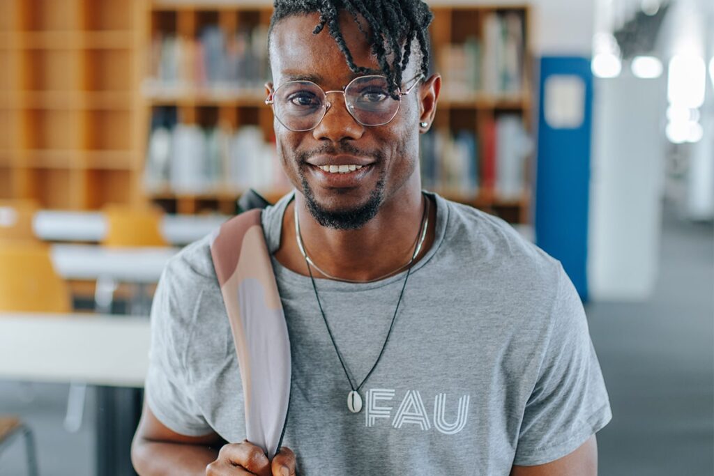 Männlich gelesene Person trägt ein graues T-Shirt mit dem Logo der Friedrich-Alexander-Universität Erlangen-Nürnberg. Im Hintergrund sieht man eine Bibliothek. Die Person trägt eine Brille, Schmuck und einen Rucksack.