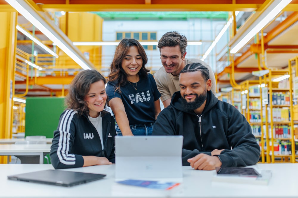 Eine Gruppe Studierender blickt gemeinsam auf auf Tablet. Im Hintergrund sieht man eine Bibliothek.