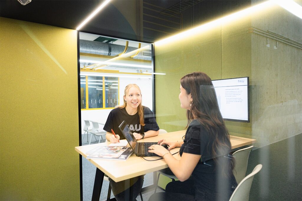 Zwei weiblich gelesene Personen sitzen in einer grünen Silent Box. Die eine Person arbeitet an einem Laptop, daneben liegt ein Magazin. Im Hintergrund sieht man einen Bildschirm, der an der Wand angebracht wurde.