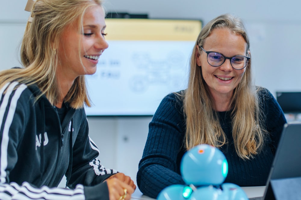 Zwei weiblich gelesene Personen sitzen vor einem Tablet zusammen. Im Hintergrund sieht man ein interaktives Whiteboard.