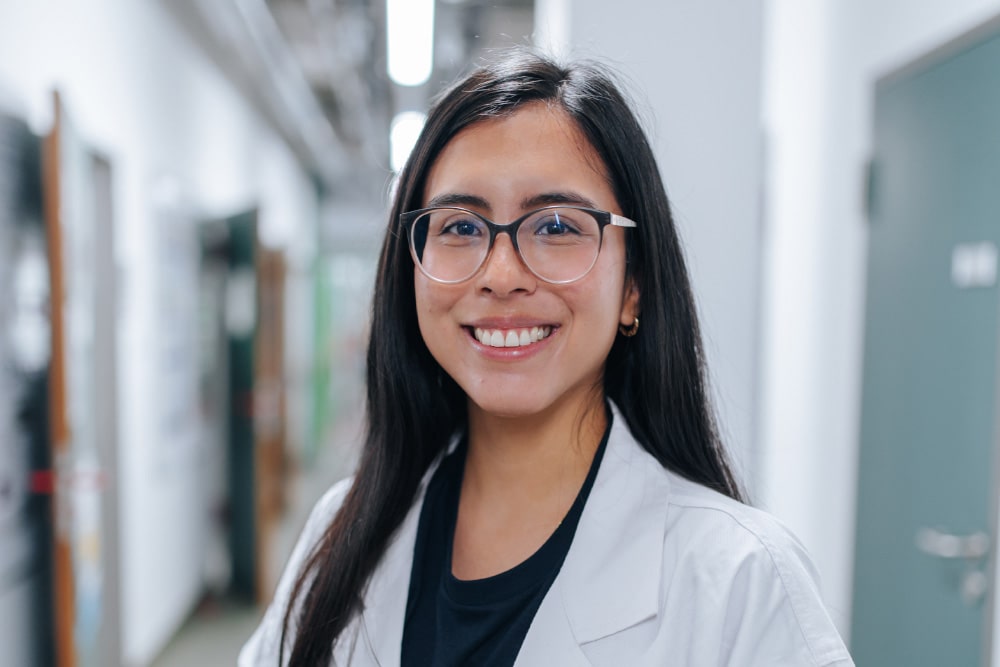 Weiblich gelesene Person mit langen schwarzen Haaren, einer Brille und in einem weißen Kittel lächelt in die Kamera.
