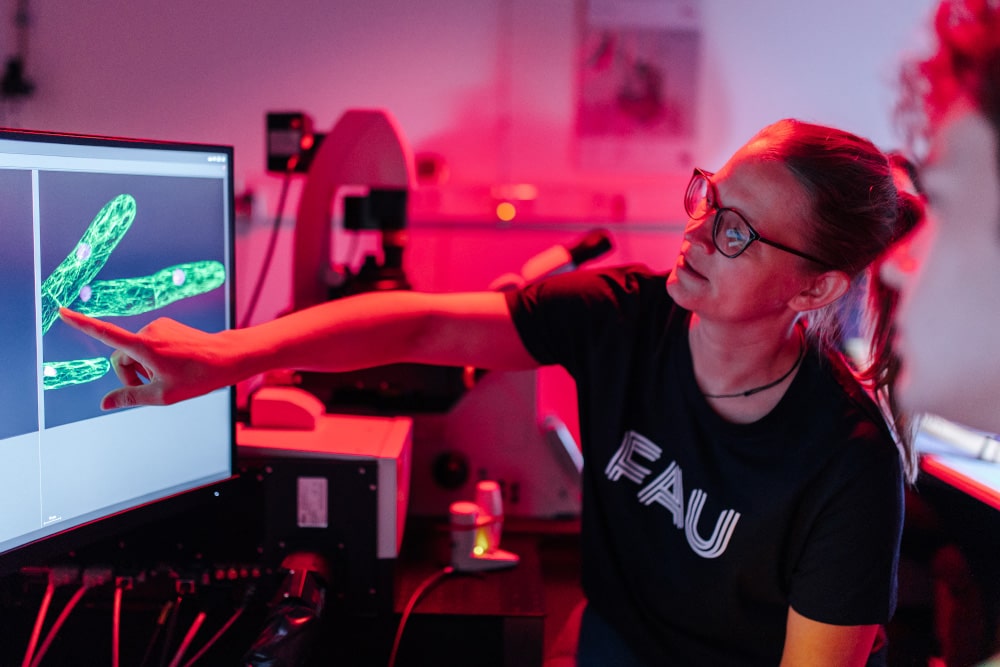 Eine weiblich gelesene Person steht in einem rot belichteten Labor und deutet auf ein Bild auf einem Bildschirm.