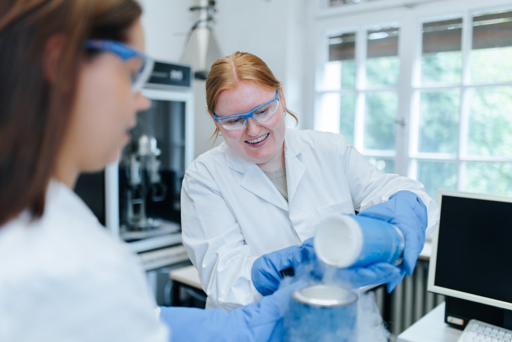 Zwei weiblich gelesene Personen stehen in einem Labor und arbeiten an einem chemischen Experiment. Beide tragen weiße Kittel, Handschuhe und Schutzbrillen.