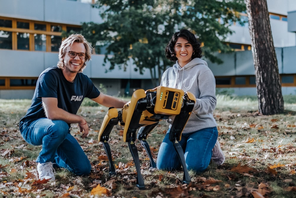 Zwei Personen knien vor einem weißen Gebäude mit einem gelb-schwarzen Roboter auf vier Beinen, der zwischen ihnen stat. Beide Personen lächeln in die Kamera.