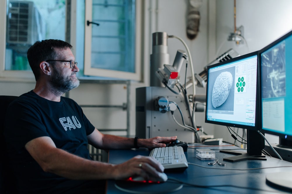 Eine männlich gelesene Person sitzt an einem Schreibtisch vor zwei Bildschirmen. Im Hintergrund sieht man Forschungsgeräte.