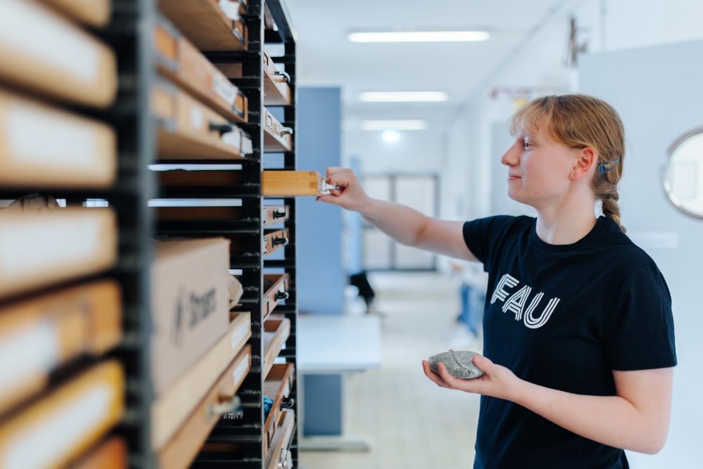 Eine weiblich gelesene Person steht vor einem Schrank mit vielen Holzschubladen, von denen sie eine gerade öffnet. In der Hand hält sie einen Stein.