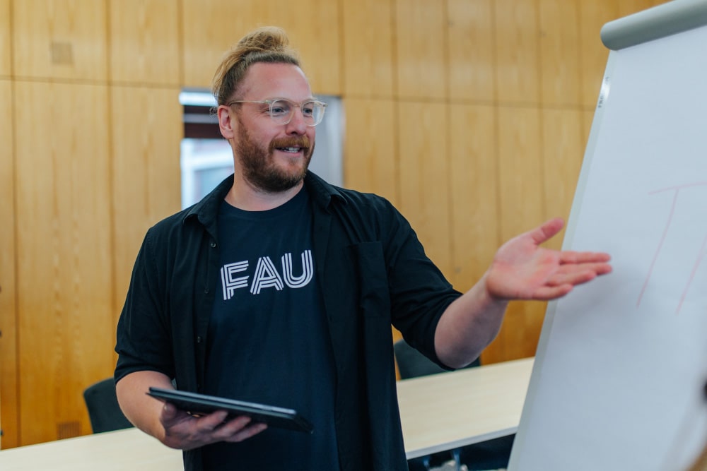 Eine männlich gelesene Person steht vor einem Flipchart, an dem sie etwas erklärt. In der Hand hält sie ein Tablet und sie trägt Brille.