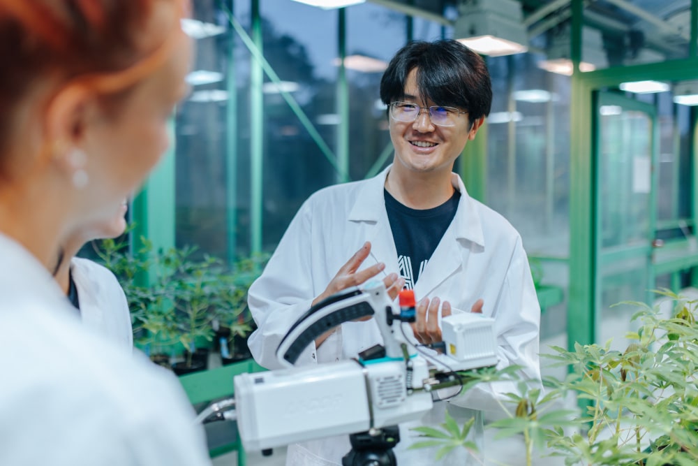 Eine Person mit asiatisch anmutenden Gesichtszügen und kurzen schwarzen Haaren steht vor einem technischen Gerät zwischen Topfpflanzen. Sie trägt einen weißen Laborkittel und eine Brille.