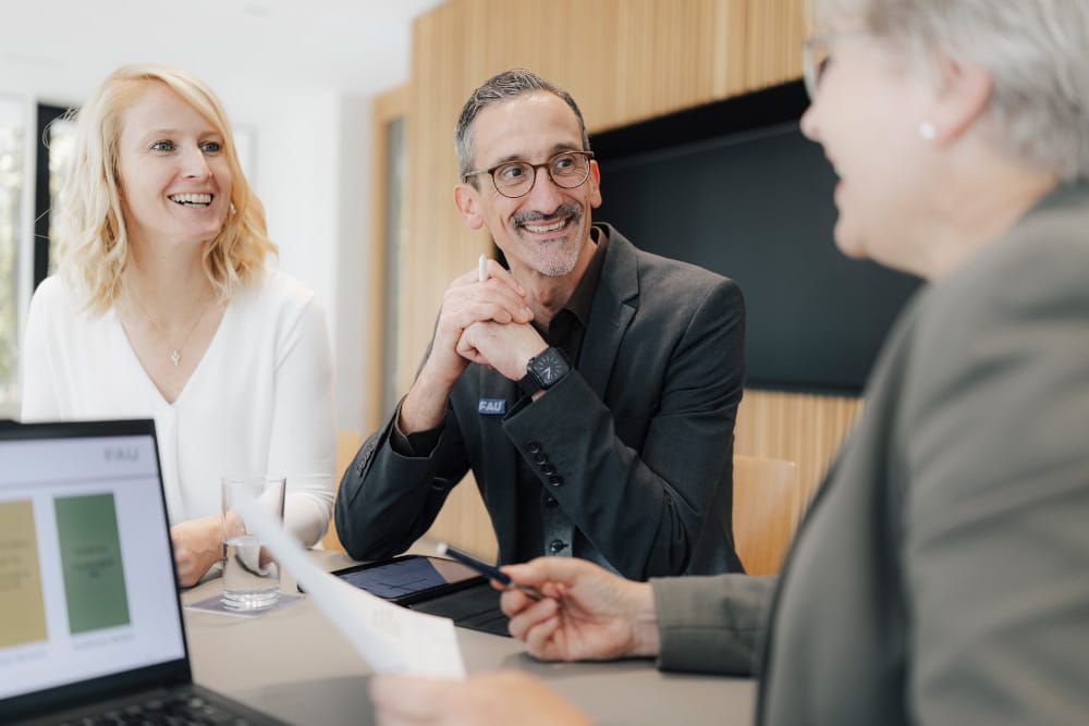 Drei Personen sitzen in einer Besprechung zusammen. Alle sind Business Casual gekleidet. Auf dem Tisch steht ein Laptop.